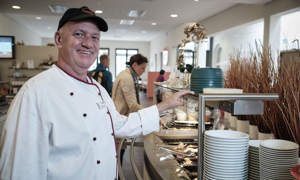 Mehrweg-System: Betriebsleiter Gerhard Böhl zeigt im Mitarbeiterrestaurant „Caruso“ auf die Re-BOWLs, die die Einwegschalen ersetzen. Neben der Einführung dieses Pfand-Systems wurde das vegetarische und vegane Speisenangebot verbessert, die Speisereste werden entweder weiterverwertet oder der städtischen Biogasanlage zugeführt. Für die Patient:innen gibt es eine vegetarische Menülinie.